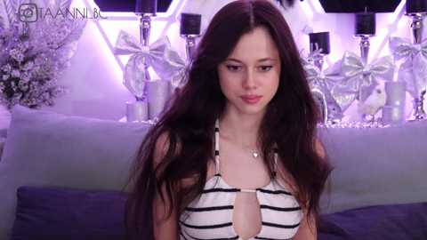 Video of a young woman with long dark hair, wearing a black and white striped halter top, sitting on a bed with a plush grey blanket. The background features a chic, modern bedroom with white and silver decor.