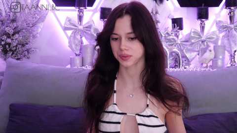 Video of a young woman with long dark hair, wearing a black-and-white striped halter top, sitting on a couch in a dimly lit room with decorative pillows and a butterfly-patterned wall.