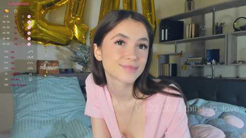 Media: A video of a young woman with long brown hair, fair skin, and a slight smile, wearing a pink top. Background features a calendar, gold balloon, bookshelf, and a bed with blue sheets.