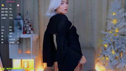 Media: A video of a slender, platinum-blonde woman with a pale complexion in a black, long-sleeved dress, posing seductively in a cozy, lit room with a lit Christmas tree and glowing candles.