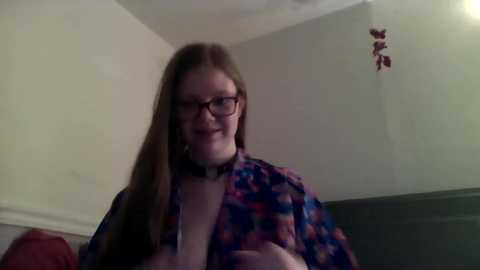 Media: Video of a smiling, fair-skinned woman with long, straight brown hair, wearing glasses, a floral blouse, and a black choker, seated in a dimly lit room with cream walls and a red cushion.