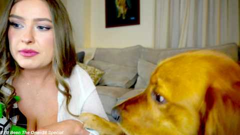 Media: Video of a fair-skinned woman with long brown hair, wearing a white blouse, sitting next to a golden retriever. The background shows a beige sofa and a framed picture on a cream-colored wall.