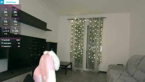 Media: A video of a minimalist, modern living room with a TV mounted on the wall, a grey couch, and a person in a pink coat standing in the foreground.