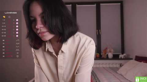 Media: Video of a young woman with shoulder-length black hair, wearing a beige button-up shirt, sitting on a bed with a white comforter. The room has a television displaying a \"LOVEING\" logo, and a window with blinds.