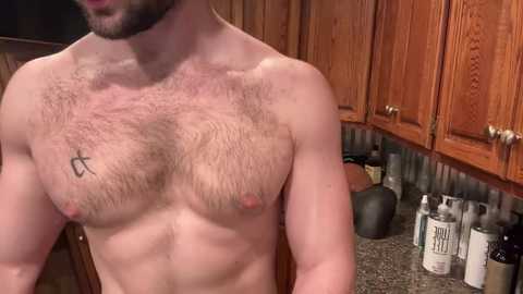 Media: Video of a shirtless man with a hairy chest, wearing a beard, standing in a kitchen with wooden cabinets and countertop.