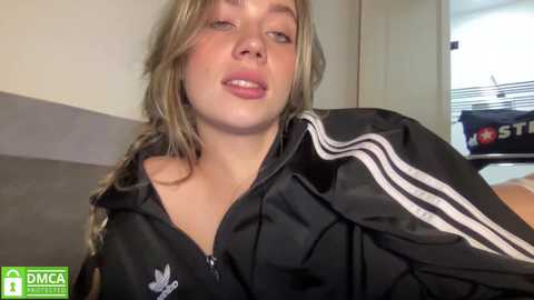 Media: Video of a blonde, fair-skinned woman with freckles, wearing a black Adidas tracksuit, in a plain room with a white door and a grey carpet.