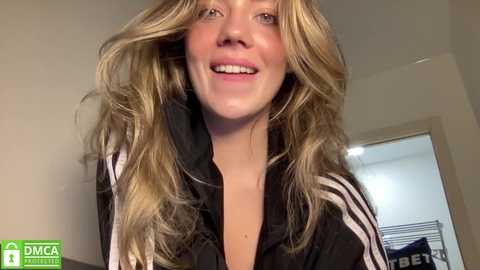 Media: Video of a smiling, light-skinned young woman with wavy, blonde hair and light makeup. She wears a black and white Adidas jacket indoors.