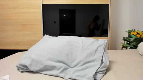 Media: Video of a neatly made bed with a white pillow and duvet, set against a minimalist bedroom with light wood furniture, a mirror, and a vase of sunflowers.