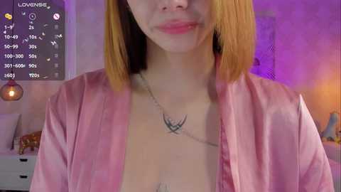 Media: A video of a young woman with fair skin, shoulder-length blonde hair, and a silver necklace featuring a small bird tattoo. She wears a pink satin robe. The background features a dimly lit, cozy bedroom with purple lighting.