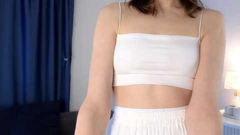 Video of a slim, light-skinned woman with brown hair, wearing a white, sleeveless crop top and matching high-waisted shorts, standing indoors against a blue curtain and a lit lamp.