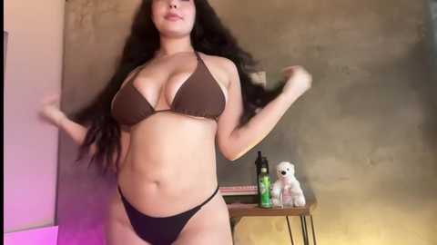 Media: Video of a curvy woman with long black hair, wearing a brown bikini, standing in a dimly lit room with concrete walls, a green bottle, and a stuffed dog on a wooden table.