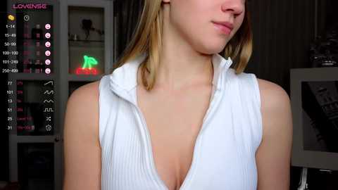 Media: A video of a fair-skinned, blonde woman wearing a white ribbed sleeveless top with a deep V-neck, showcasing her cleavage. She stands in a dimly lit room with a TV displaying a live stream.