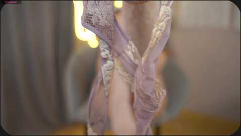 Media: Video of a close-up of a woman's legs in light purple lace panties, hands pulling them down. Background is blurred, showing a dimly lit room with a yellow light.