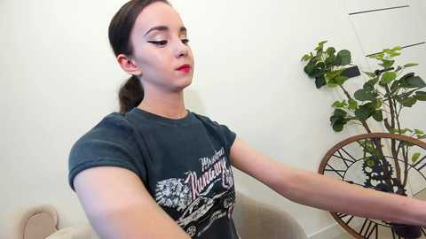 Media: Video of a fair-skinned woman with brown hair in a ponytail, wearing a dark T-shirt with white graphic text, sitting in a beige armchair in a minimalist room with a green plant.