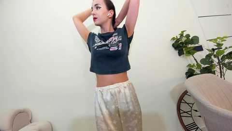 Media: Video of a slim, fair-skinned woman with brown hair, wearing a dark T-shirt and light pajama pants, stretching against a plain white wall.