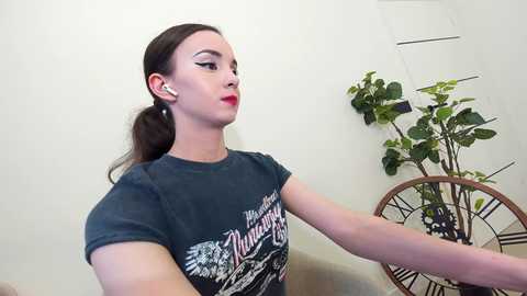 Media: Video of a fair-skinned woman with dark hair, red lipstick, wearing headphones, dark T-shirt with vintage design, seated indoors next to a potted plant and clock on a white wall.