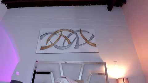 Media: Video of a modern room with a white wall featuring a large abstract painting of intertwining golden and gray lines. Below, white shelves hold various items, including a lamp emitting a warm glow, and a purple light source is visible on the left.