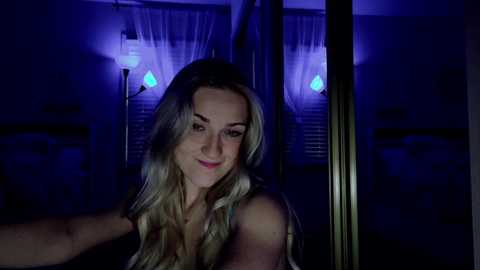 Media: A video of a blonde woman with fair skin and a slight smile, wearing a sleeveless top, standing in front of a window with blue light from behind, dimly lit room in the background.