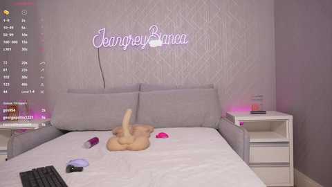 Media: Video of a modern, minimalist bedroom with a gray upholstered headboard, a plush white bed, a pink dildo on the bed, a keyboard on the left, and a pink lamp on the right nightstand. The wall features a pink neon sign reading \"Jeangrey Banks.\