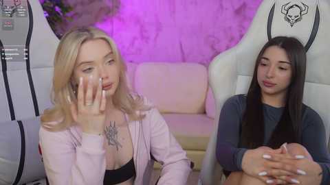 Media: Video of two women: blonde with pink jacket, black bra, and tattoo; brunette in grey top, holding hands. Background: white chair, purple wall.