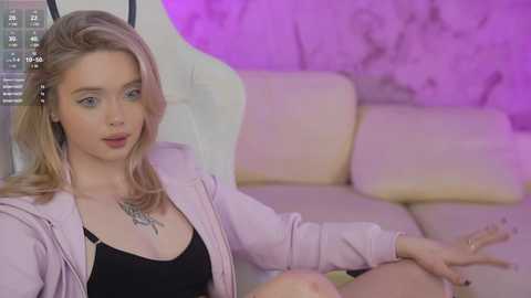 Media: Video of a blonde Caucasian woman with fair skin, wearing a pink blazer and black top, sitting on a white leather couch. Background features a pink wall with a modern abstract design.