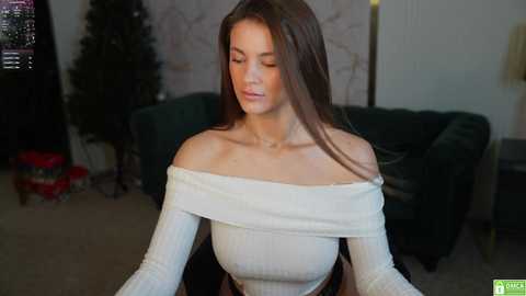 Media: Video of a fair-skinned woman with long brown hair, wearing an off-shoulder white sweater, sitting in a dimly lit living room with a dark green couch, Christmas tree, and presents in the background.