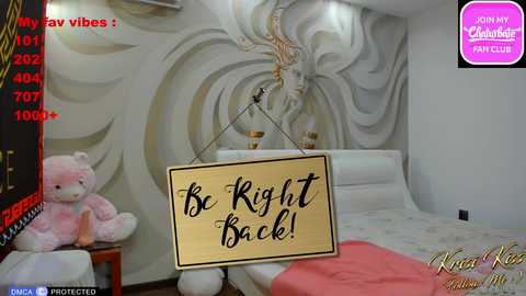 Media: Video of a bedroom with a white bed, beige walls with gold accents, a stuffed animal, and a sign reading \"Be Right Back!\" in a playful font.