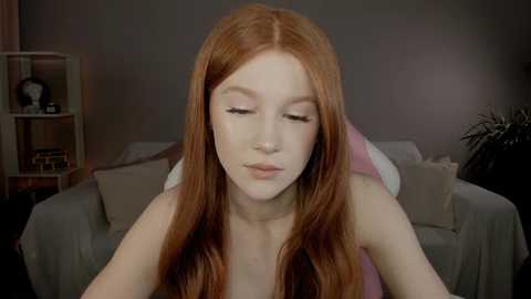 Media: Video of a fair-skinned, red-haired woman with long hair, wearing a pink top, sitting on a beige sofa with a gray blanket, in a dimly lit room with a shelf in the background.