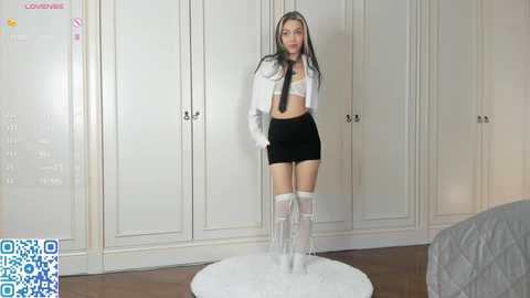 Media: Video of a slender, fair-skinned woman with long black hair, wearing a white cropped jacket, black mini skirt, white lace bra, and thigh-high white boots, standing in a minimalist bedroom with white paneled wardrobe.