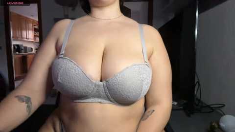 Video of a woman in a light gray lace bra, showing ample cleavage, with a tattoo on her left forearm, in a dimly lit room with kitchen cabinets and a microwave in the background.