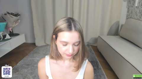 Video of a young Caucasian woman with shoulder-length blonde hair, wearing a white tank top, seated on a gray rug in a modern living room with beige curtains and a white couch.