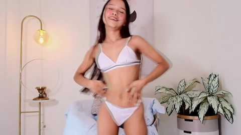 Media: A video of an Asian woman with long dark hair, wearing a white bra and panties, smiling, standing in a minimalist room with a plant, lamp, and bed.