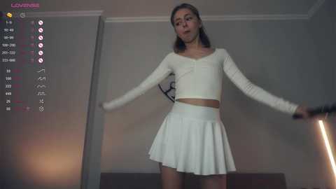 Media: Video of a slender, light-skinned Asian woman with shoulder-length brown hair, wearing a white, long-sleeved crop top and short pleated skirt, standing confidently indoors.