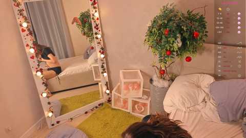 Video of a cozy, well-lit bedroom with a white bed, green rug, and large mirror adorned with red and white lights. A potted plant with red and white ornaments stands beside a white nightstand with decorative boxes.