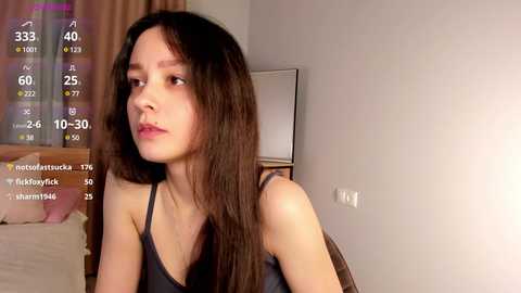 Media: Video of a young woman with long dark hair, light skin, and small breasts, wearing a black tank top, sitting indoors by a window with a digital clock displaying 24:6:30.