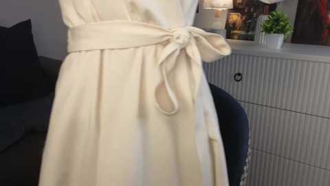 Media: Video of a woman in a cream-colored dress with a large bow at the waist, standing in front of a gray, textured cabinet with a potted plant and a lit lamp on top.