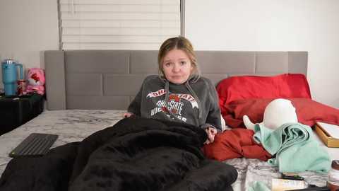 Media: Video of a young Caucasian girl with light brown hair, wearing a gray sweatshirt and black pants, lying on a bed with red and white bedding, surrounded by clothes and a stuffed animal.