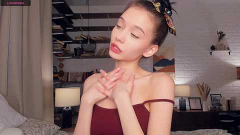 Media: Video of an Asian woman with light skin, straight brown hair, wearing a burgundy off-shoulder top, sitting on a bed in a cozy, modern loft apartment with exposed brick walls, shelves, and a lamp.