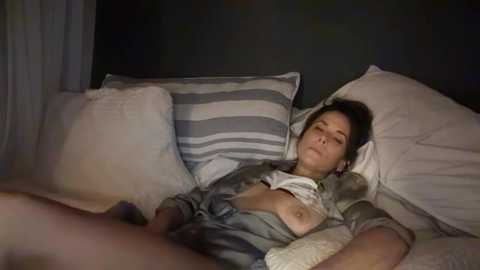 Media: A video of a woman with medium breasts, lying on a bed with white and grey striped pillows, wearing a grey shirt, partially unbuttoned.