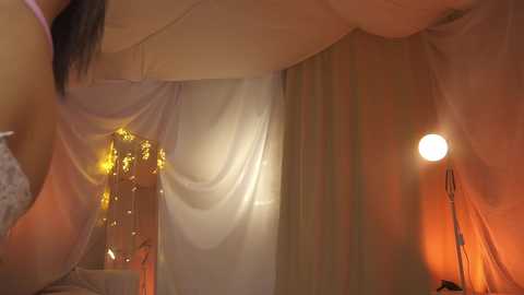 Media: Video of a woman with long dark hair, partially covered by sheer white drapes, standing in an intimate, dimly lit room with a warm orange lamp and golden string lights.