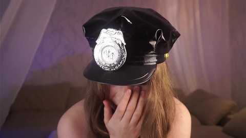 Media: Video of a woman wearing a black police cap, partially covering her face with her hand, in a dimly lit room with sheer curtains and a brown couch.