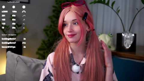 Media: Video of a smiling, fair-skinned woman with long, pink hair and pink sunglasses, sitting on a sofa in a modern, dimly lit living room.