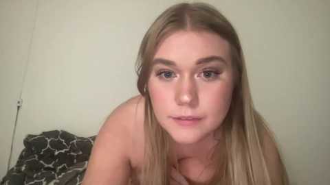 Media: Video of a young, light-skinned, blonde woman with blue eyes, wearing minimal makeup, lying on a bed with a patterned pillow. She has a soft expression and is topless, with a plain, light-colored wall background.