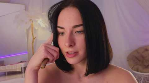 Video of a fair-skinned, dark-haired woman with a medium build, light makeup, and straight shoulder-length hair. She's topless, holding her chin, in a softly lit, white room with a plant in the background.