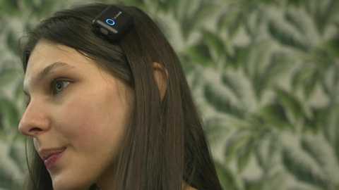 Media: Video of a young woman with straight, dark brown hair, wearing a small, black hair clip with a blue logo. She has fair skin and is smiling. Background is out of focus, featuring green foliage.