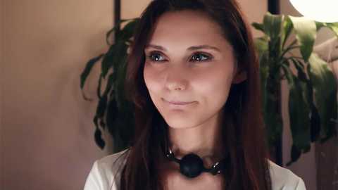 Media: Video of a young woman with long brown hair, wearing a white shirt and a black choker, standing indoors with a green plant and beige wall in the background.