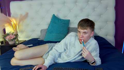 Media: Video of a young man lying on a bed, wearing a white sweater, eating a popsicle, with a teal pillow, and a purple wall in the background.
