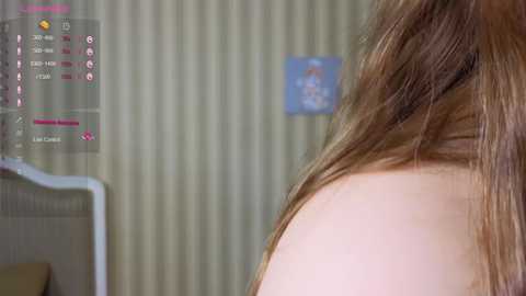 Media: Video of a close-up of a woman's shoulder and back, with light brown hair, against a beige, vertical-striped wall. In the background, there's a digital interface displaying health metrics, suggesting a medical context.