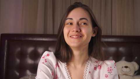 Media: Video of a young woman with straight brown hair, wearing a white floral dress, smiling in a cozy bedroom with a dark brown leather headboard.