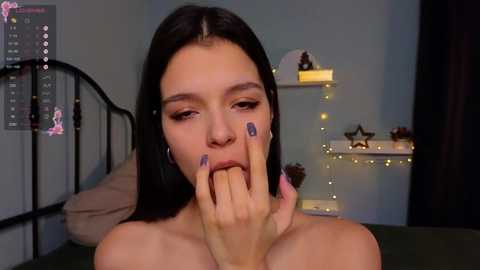 Video of a young woman with fair skin and long black hair, wearing purple nail polish, seductively licking her fingers in a dimly lit bedroom with a black metal bed and fairy lights.